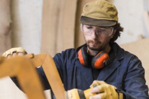 close up male carpenter wearing safety glasses ear defender around his neck using electric sander 23 2147944827