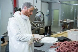 man wearing white coat hairnet reviewing clipboard meat plant near raw sausages machinery 13339 390360